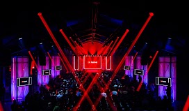 An auditorium with people seated. At the front, a black screen with the YouTube icon and the word “Festival” on it. Six screens, three on either side of the people, are on the walls with the same logo and copy. Red strobe lights shine over the audience.