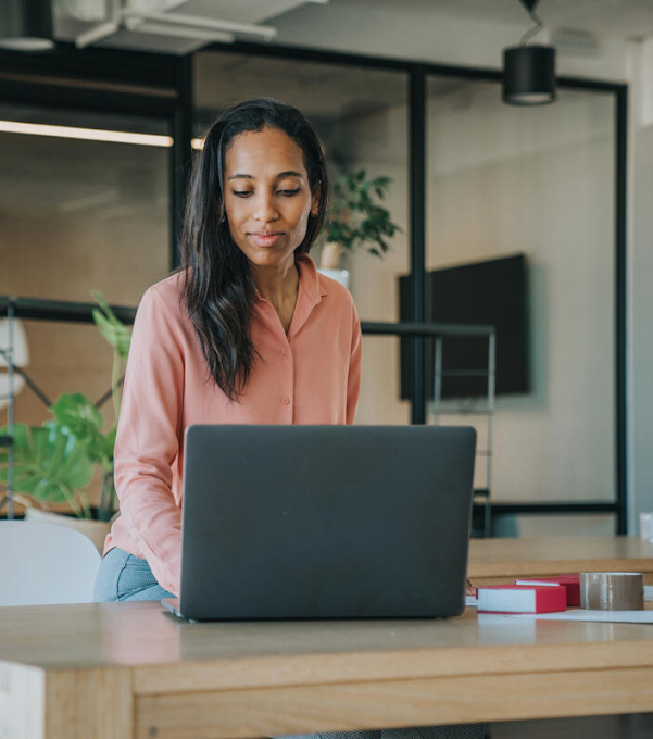 A woman is sitting at a table, looking at her laptop. There are indoor plants on a shelf in the background.