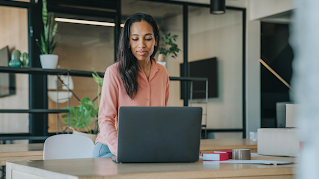 A woman is sitting at a table, looking at her laptop. There are indoor plants on a shelf in the background.