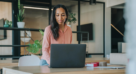 A woman is sitting at a table, looking at her laptop. There are indoor plants on a shelf in the background.