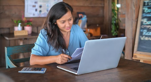 A woman is sitting at a table, holding papers and a pen. A laptop and a calculator can be found on the table in front of her.