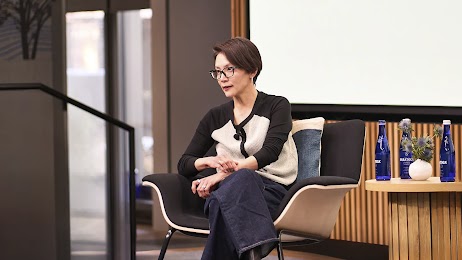 Elissa Lee, senior director of media measurement and optimization at Google, sits in a chair and speaks on a panel. Lee has light skin, short dark hair, and wears black glasses, a cream and black sweater, and black pants.