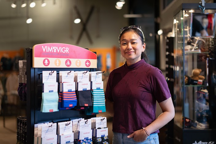 Michelle Huie, Founder & Owner of VIM & VIGR, stands next to a display rack filled with compression legwear, smiling at the camera in a retail setting.