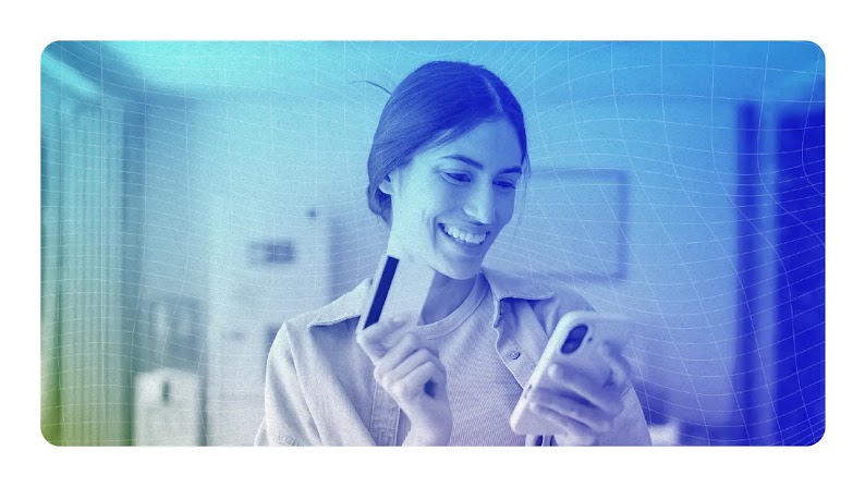 Mulher de cabelos escuros sorrindo e fazendo uma transação no celular, com o cartão em mãos.