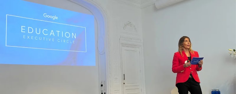 Celia Villalobos vestida con una chaqueta roja y pantalones negros está de pie en un escenario, hablando en un evento. Detrás de ella, una pantalla azul proyecta Google EDUCATION EXECUTIVE CIRCLE