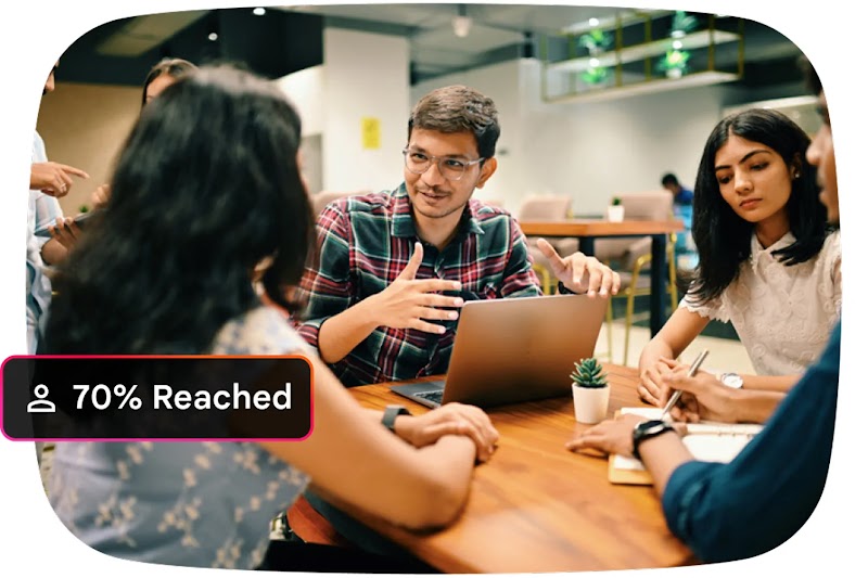A group of young professionals collaborates around a wooden table with a laptop and notebooks in a modern office. A black text overlay in the corner reads: "70% Reached”.