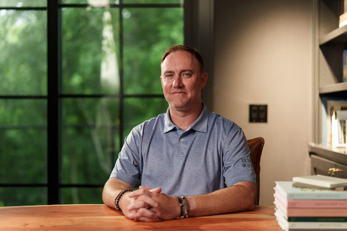 Garrett Peters, Co-Founder and CEO of Duncan & Stone Paper Co., sits at a wooden desk with his hands clasped, wearing a blue polo shirt, in front of a window overlooking green trees.