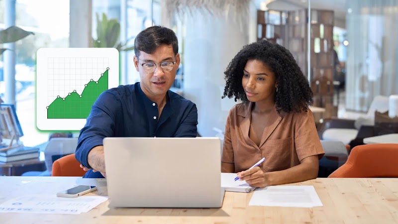 A medium shot of a man wearing glasses and a dark blue shirt and a woman with dark curly hair wearing a brown button-down shirt sitting at a desk and looking at a laptop in a modern office with natural light. To the left is an icon of a green graph with white lines.