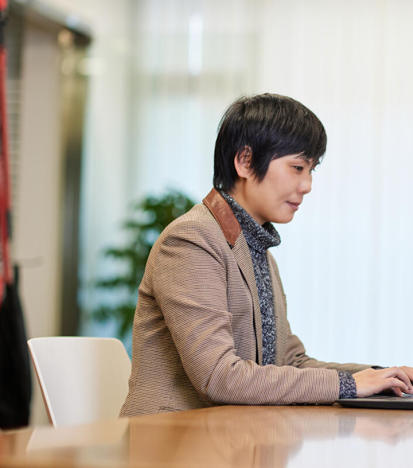 A woman photographed from the side is sitting at a table and typing on a laptop.