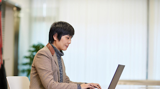 A woman photographed from the side is sitting at a table and typing on a laptop.