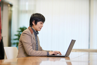 A woman photographed from the side is sitting at a table and typing on a laptop.