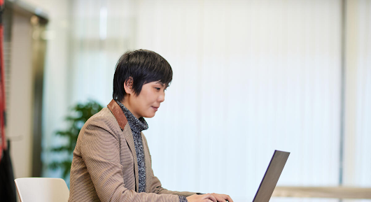 A woman photographed from the side is sitting at a table and typing on a laptop.