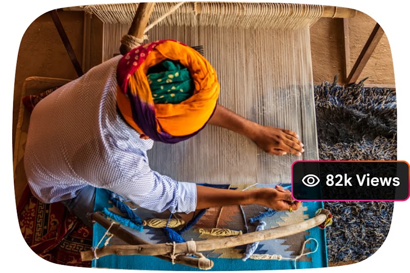 An overhead view of a person wearing a colorful orange and green turban weaving a textile on a traditional wooden loom. A black text overlay in the corner reads: "82k Views”.