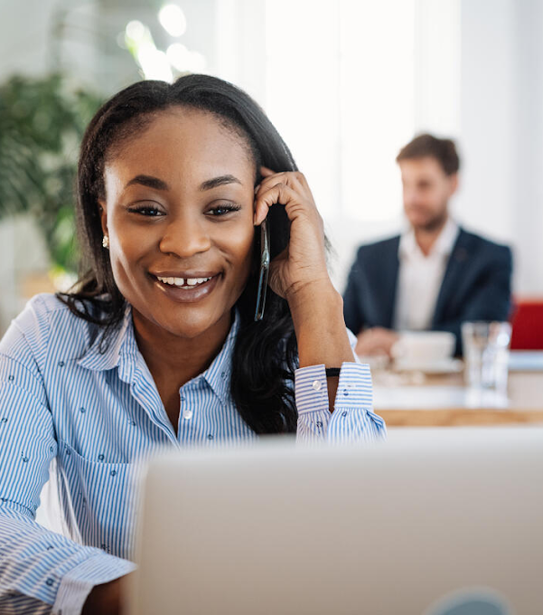 A woman is talking on the phone smiling, while looking at her laptop. There is a plant and a man working in the blurred background.