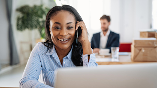A woman is talking on the phone smiling, while looking at her laptop. There is a plant and a man working in the blurred background.