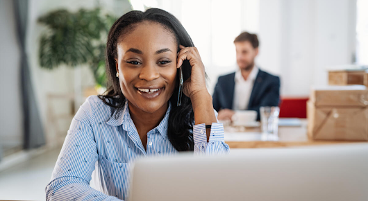 A woman is talking on the phone smiling, while looking at her laptop. There is a plant and a man working in the blurred background.