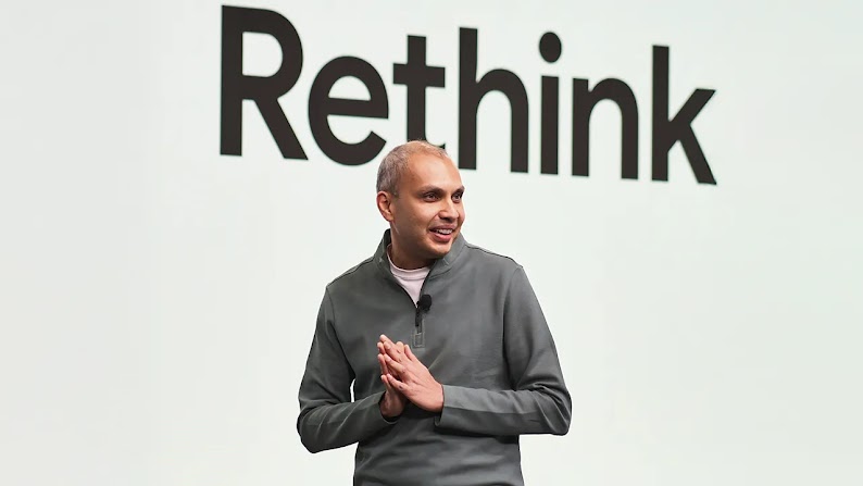 Gaurav Bhaya, VP and GM of buying, analytics, and measurement at Google gives a talk in front of a digital screen reading “Rethink.” Bhaya has short, light  hair, medium skin, and wears a gray zip-neck pullover over a white t-shirt.