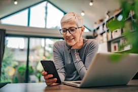 A middle-aged woman with short blonde hair and glasses smiles while looking at her smartphone. She sits at a desk with an open laptop in a bright, modern room with large windows and high ceilings.