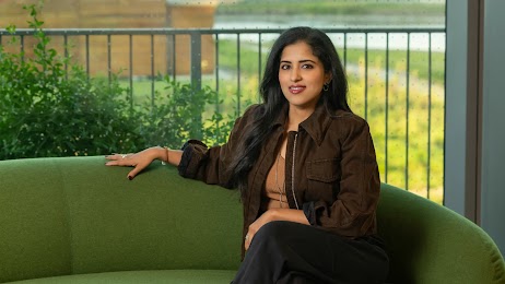 Vidhya Srinivasan, VP and GM of Ads and Commerce at Google, sits on a green couch in front of a window overlooking a shrub and patio rail. Srinivasan has medium skin, long dark hair, and wears a casual brown jacket over a tan top with black slacks.