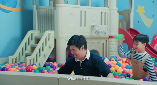A frustrated man pulling his hair in a colorful ball pit while a young boy winds up to throw a plastic ball at him, with the subtitle "Whats is it?"