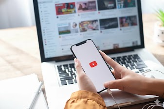 A close-up of hands holding a modern smartphone displaying the YouTube app logo, with a laptop showing the YouTube website feed open in the background.