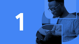 A person with dark skin, short hair, and glasses sits cross-legged while typing on a laptop.