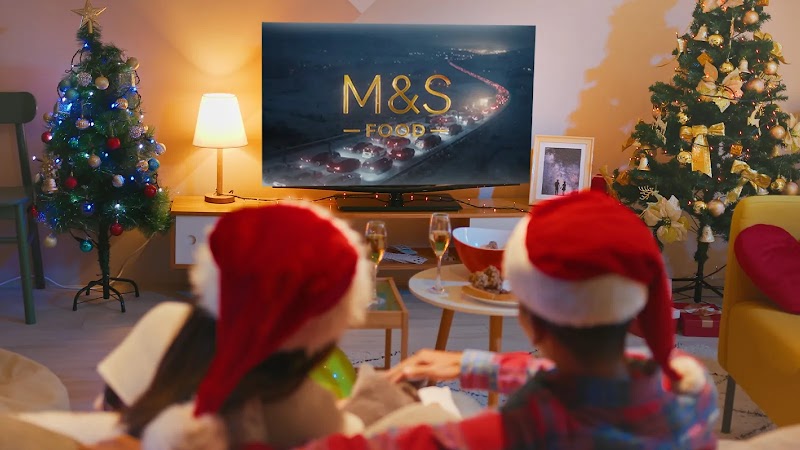 Two people sit on a sofa facing a TV. On the screen, an ad is playing with the copy “M&S FOOD” in the centre. The room is decorated with two Christmas trees, one either side of the TV. On the table in front of the couple, two champagne glasses.