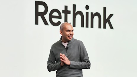 Gaurav Bhaya, VP and GM of buying, analytics, and measurement at Google gives a talk in front of a digital screen reading “Rethink.” Bhaya has short, light  hair, medium skin, and wears a gray zip-neck pullover over a white t-shirt.