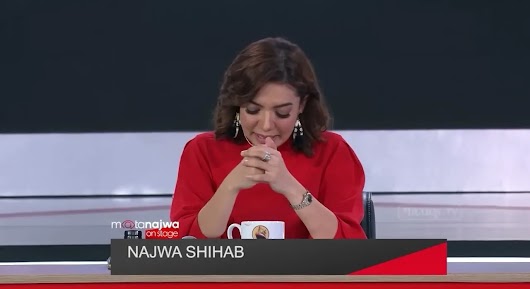 A video screenshot of journalist Najwa Shihab seated at a broadcast desk, wearing a red shirt and clasping her hands together with a thoughtful expression.
