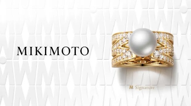 Mikimoto 高端珠寶品牌視覺示意。白色紋理背景搭配「MIKIMOTO」品牌名稱，展示金色鑲鑽珍珠戒指，中央白色珍珠搭配金色戒身，密鑲鑽石呈放射狀排列。下方標註「M Signature」珍珠系列標誌，代表品牌經典珠寶工藝與奢華設計。