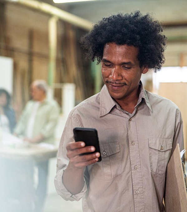 A man wearing a grey long sleeve shirt is standing and looking at his phone, smiling. There are people behind him in the blurred background.