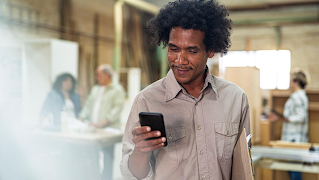 A man wearing a grey long sleeve shirt is standing and looking at his phone, smiling. There are people behind him in the blurred background.