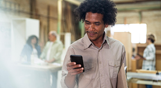 A man wearing a grey long sleeve shirt is standing and looking at his phone, smiling. There are people behind him in the blurred background.