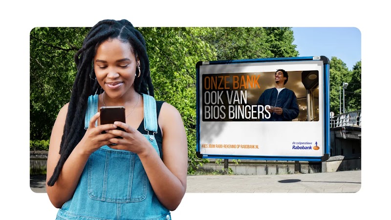 Woman with dreadlocks using a smartphone outdoors, with a street billboard behind her displaying the text "ONZE BANK OOK VAN BIOS BINGERS" next to the Rabobank logo. 