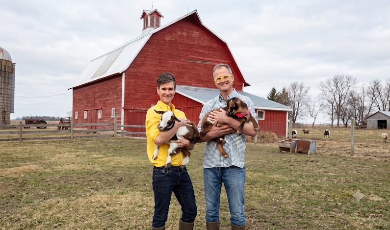 Les fondateurs de Beekman 1802, Josh Kilmer-Purcell et Brent Ridge, sourient à l’appareil photo alors qu’ils se tiennent debout devant une grange avec chacun une chèvre dans les bras.’.