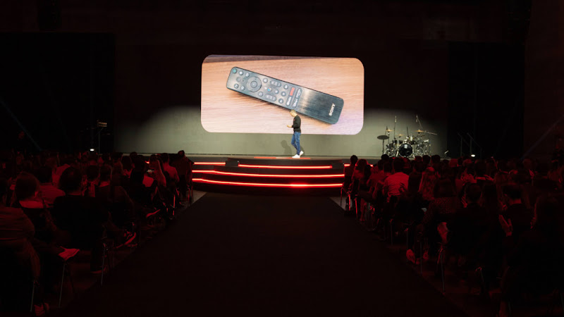 Sobre el escenario se proyecta la imagen de un mando a distancia con un botón de YouTube, Javi Vítores con jersey negro y vaqueros, camina hacia la izquierda mientras el público le observa desde las butacas.