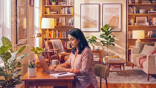 A woman in a patterned pink kurta sitting at a wooden desk working on a laptop in a warmly lit, plant-filled living room with bookshelves.