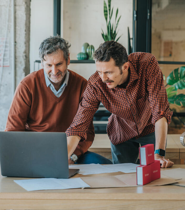 Two men in front of a laptop. The man on the right is pointing at the screen and talking as the man on the left listens.
