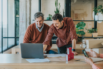 Two men in front of a laptop. The man on the right is pointing at the screen and talking as the man on the left listens.