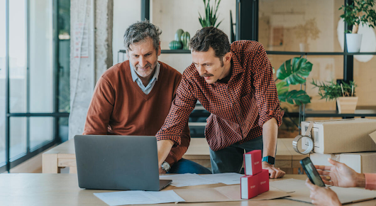Two men in front of a laptop. The man on the right is pointing at the screen and talking as the man on the left listens.