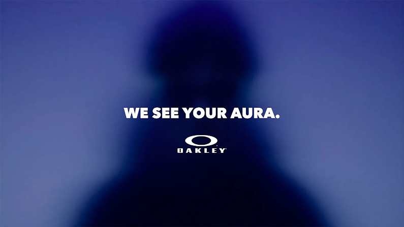 Blurry silhouette of a person against a hazy blue and purple background. White text centered on the image reads: "WE SEE YOUR AURA." with the white Oakley logo positioned directly underneath.