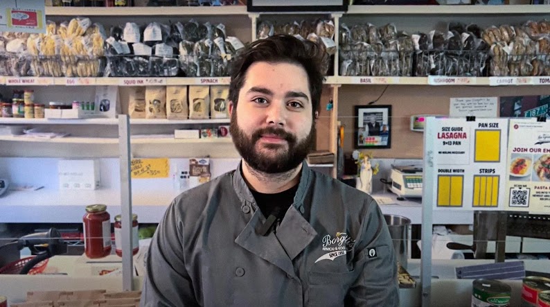 Chris Borgatti stands behind the counter of his Bronx, New York, ravioli shop. Behind him are shelves of handmade ravioli. Borgatti appears to be a young man with dark brown hair, a beard, light skin, and brown eyes. He wears a dark green chef’s coat.