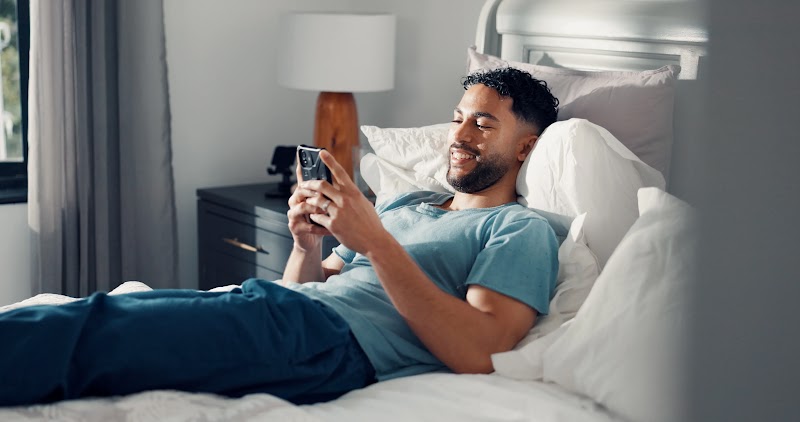 A smiling man with short curly hair and a beard lies propped up on white pillows in bed, holding a smartphone in both hands. He is wearing a blue t-shirt and dark pants in a brightly lit, modern bedroom.
