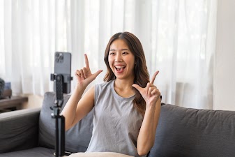 A female video blogger or influencer actively filming herself for social media, making a hand gesture and looking directly at the camera on her phone.