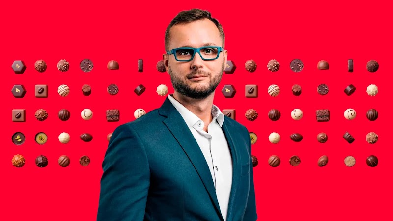 Maciej Kluk, stands against a bright red background patterned with various chocolates.