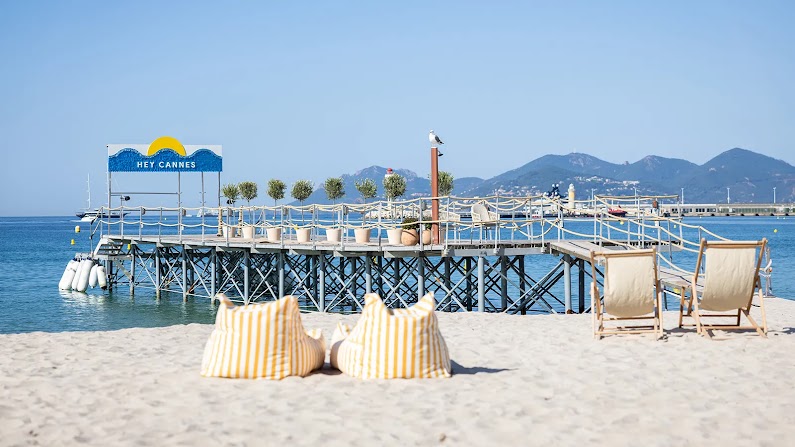 Yellow-striped beach chairs sit in the sand near a pier on a beach in Cannes, France, overlooking blue water with a clear sky and mountains in the distance.
