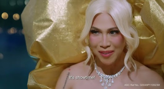 The video thumbnail shows a close-up of a person with light skin wearing a platinum blonde wig and heavy makeup, resembling a drag queen or performer. They are wearing a sparkling diamond necklace and are seated in a large, shiny golden prop or setting. The text overlay reads, "It's showtime!"