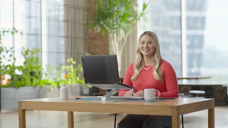 A blonde woman in a coral pink sweater smiles at the camera while sitting at a desk with a laptop and coffee mug. 