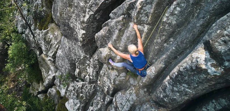 Una mujer deportista con el pelo gris escala una montaña gris y rocosa. Lleva equipo de escalada y cuerdas. Hay árboles debajo de ella, en la base de la montaña.