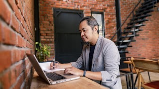 A man photographed from the side is sitting at a table, scrolling on his laptop with one hand, and taking notes in a notebook with the other.